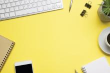 A keyboard, notebook, smartphone, pen, binder clips, plant, coffee mug, and notebook are on a bright yellow background