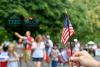 A white hand on the lower right corner of the frame, holding a small American flag. The background is blurred, but you can see a group of people standing under big green tree leaves. The TREC-TALCB logos are in the foreground at the top left corner.